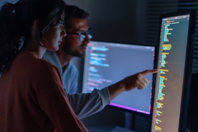 A man and woman working at a computer workstation, man pointing at code displayed on a monitor.