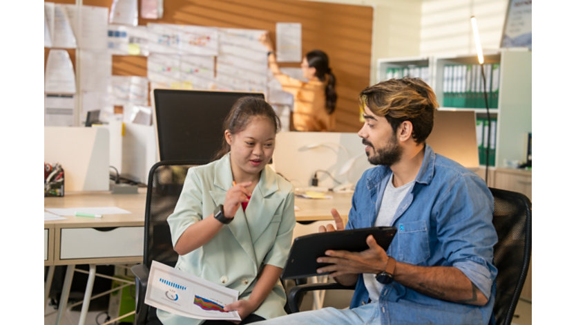 Two colleagues seated in an office collaborate on a project using a tablet. One person gestures while holding a report with charts, and the other follows along. Additional team members and planning materials are visible in the background.