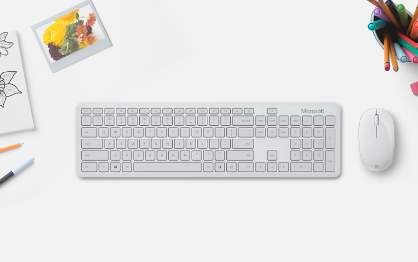Microsoft Bluetooth Keyboard junto a un Microsoft Bluetooth Mouse sobre una mesa con un lápiz, un rotulador, una goma de borrar y un cuaderno de dibujo.