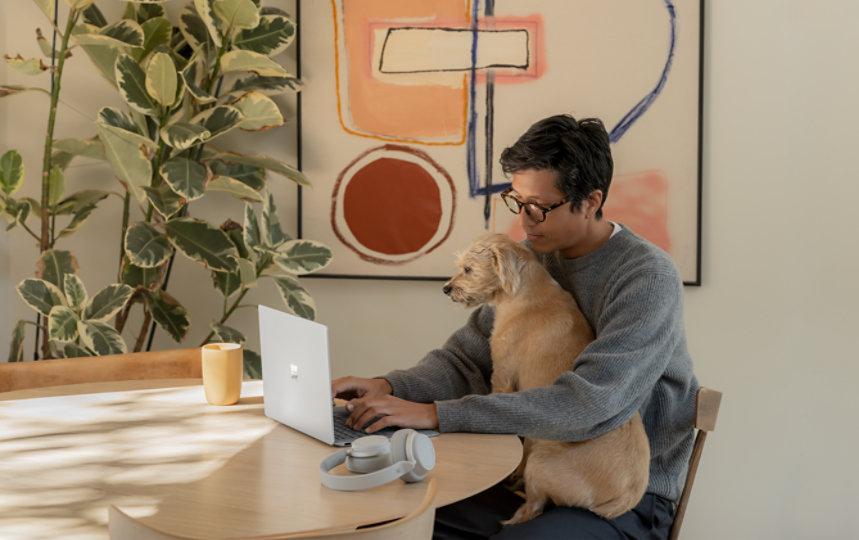 A person wearing glasses with a dog on his lap using Office home and business on a surface laptop with surface headphones nearby.