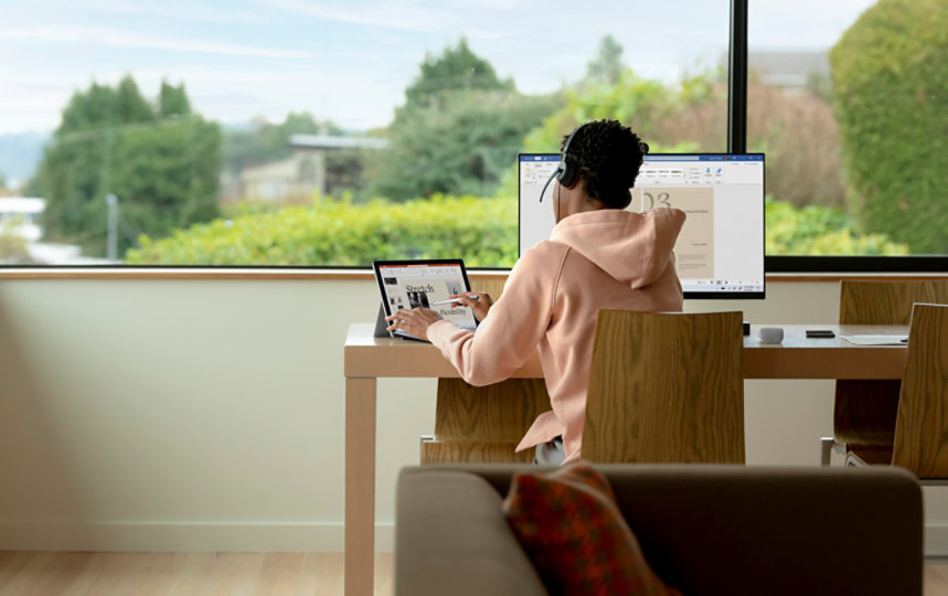 Un student folosește aplicații Office pe un laptop și un monitor, stând la birou.