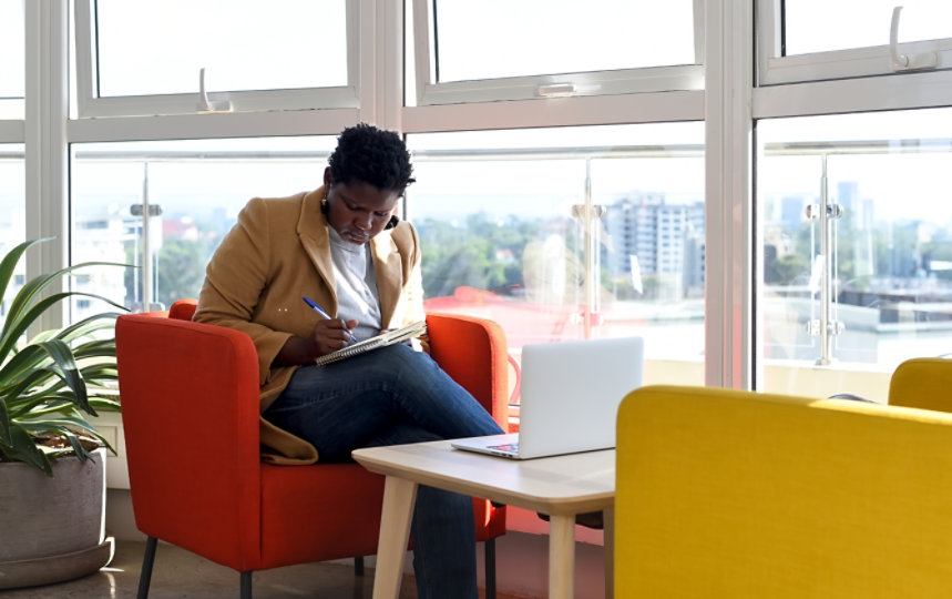 Eine Frau sitzt mit ihrem Laptop in einem modernen Büro und macht sich Notizen.