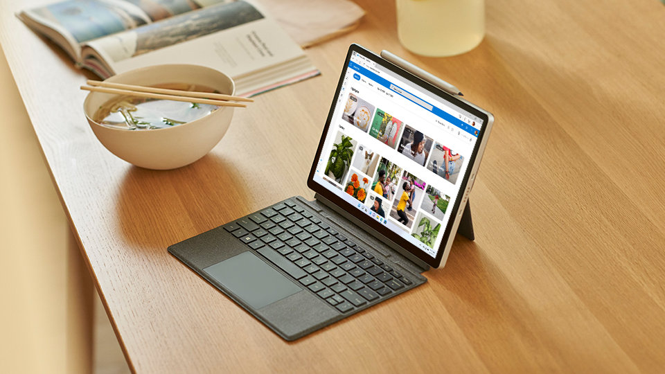 A tablet with a keyboard displaying Microsoft Edge at a table near a bowl of noodle soup and an open book.