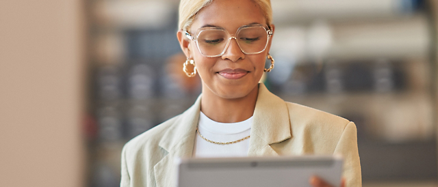 Eine Frau mit Brille und Ohrringen, die eine helle Jacke trägt, liest Inhalte auf einem Tablet mit einem konzentrierten Ausdruck.