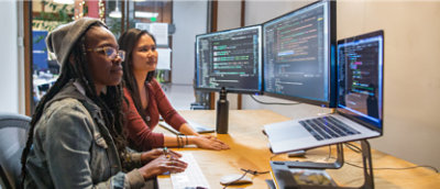 Kodların gösterildiği birden fazla ekran bulunan bilgisayar iş istasyonunda birlikte çalışan iki kişi.