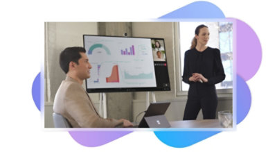 Intervenant debout dans une salle de réunion avec un grand écran affichant une présentation, un appel vidéo Teams et un collègue écoutant l’intervenant.