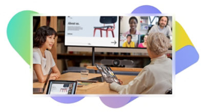 Deux personnes en réunion avec de grands écrans affichant un appel vidéo Teams et une présentation, l’une parle et l’autre tient une tablette.