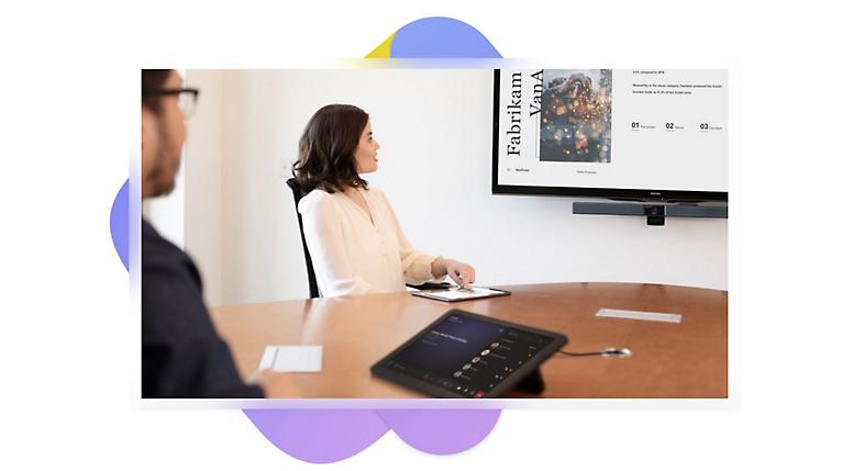 Dos personas en una sala de reuniones con un dispositivo de reunión en la mesa que muestra una llamada de Teams en curso y una presentación en un televisor