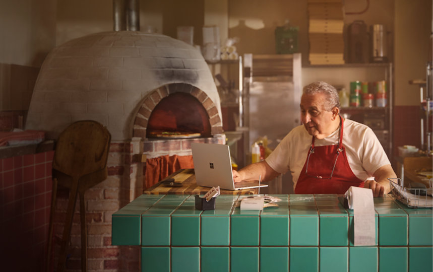 Man sitting at a countertop in a restaurant kitchen using a Surface Laptop and a calculator.