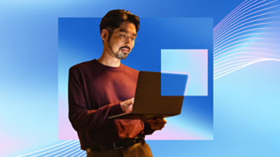 A man, standing, holding a laptop in his left hand, and using his right hand to type and operate the trackpad.