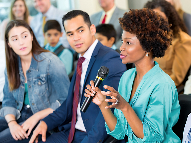 Eine Frau spricht während einer Diskussion auf einer Konferenz in ein Mikrofon, während sich aufmerksame Zuschauer im Hintergrund befinden.