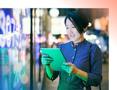 ผู้หญิงผมสั้นกําลังใช้แท็บเล็ต ยืนอยู่ข้างจอแสดงผลดิจิทัลที่มีแสงสว่างสดใสในเมืองยามค่ําคืน