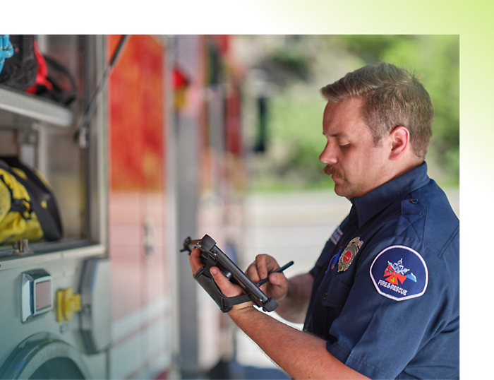 A firefighter in a blue uniform with a badge writes on a tablet beside a fire truck.