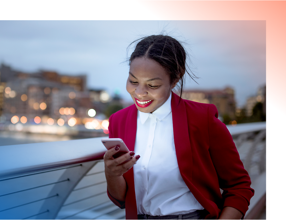A smiling woman in a red blazer using a smartphone on a city bridge at dusk.