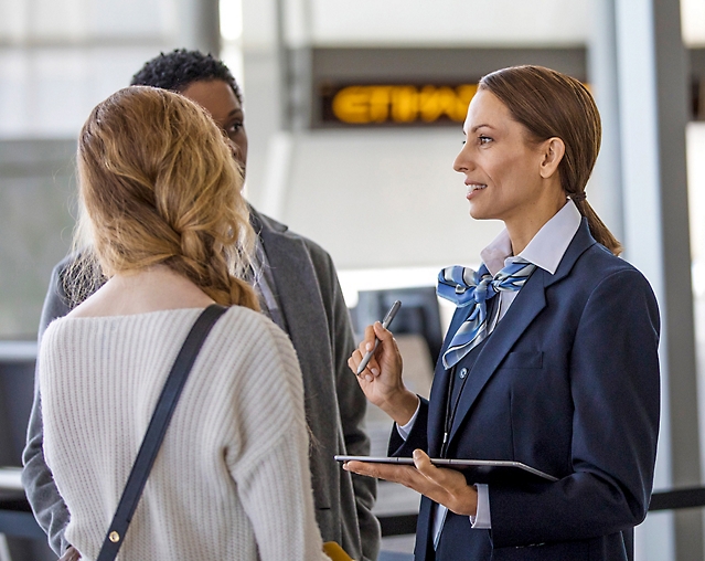 Sieviete uzvalkā sarunājas ar cilvēku grupu lidostā.