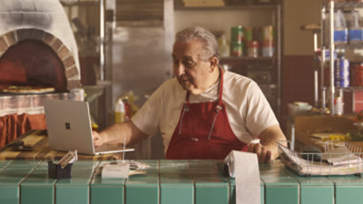 Man sitting at a countertop in a restaurant kitchen using a Surface Laptop and a calculator.