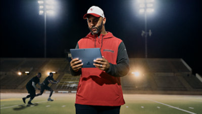 A coach uses a Surface Pro on the practice field while players run in the background.