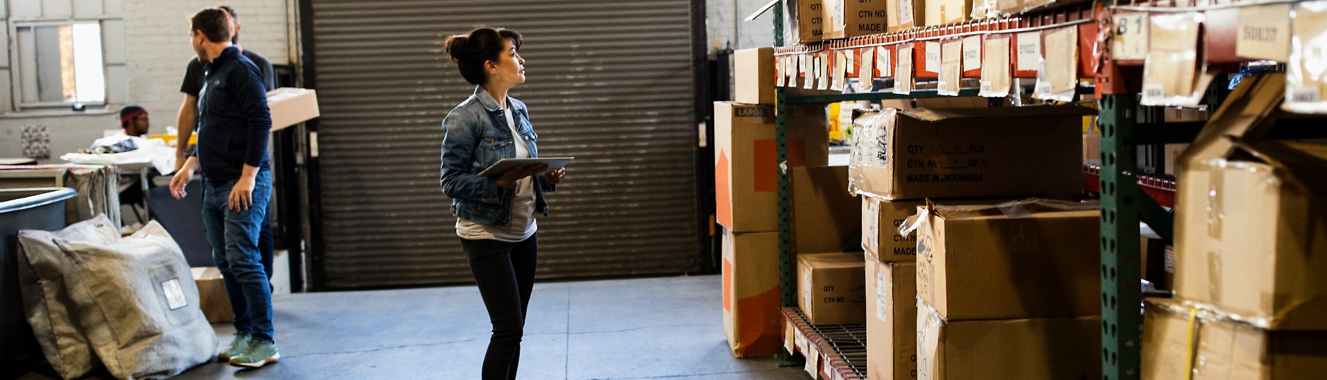 A person holding a tablet device, looking at boxes on a large shelf in a warehouse.