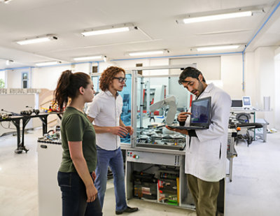 Three people are sitting at a table in a well-lit room with large windows. One person is using a laptop, another is wearing headphones, and the third person is smiling and talking.