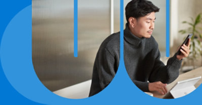 Человек сидит за столом с лежащим перед ним планшетом и смотрит на экран мобильного телефона.