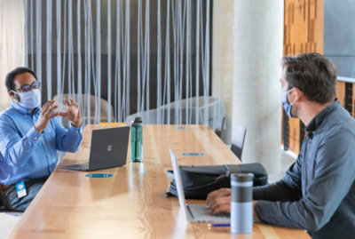 Dos hombres, con máscaras faciales, están sentados en una mesa de madera larga con portátiles. 