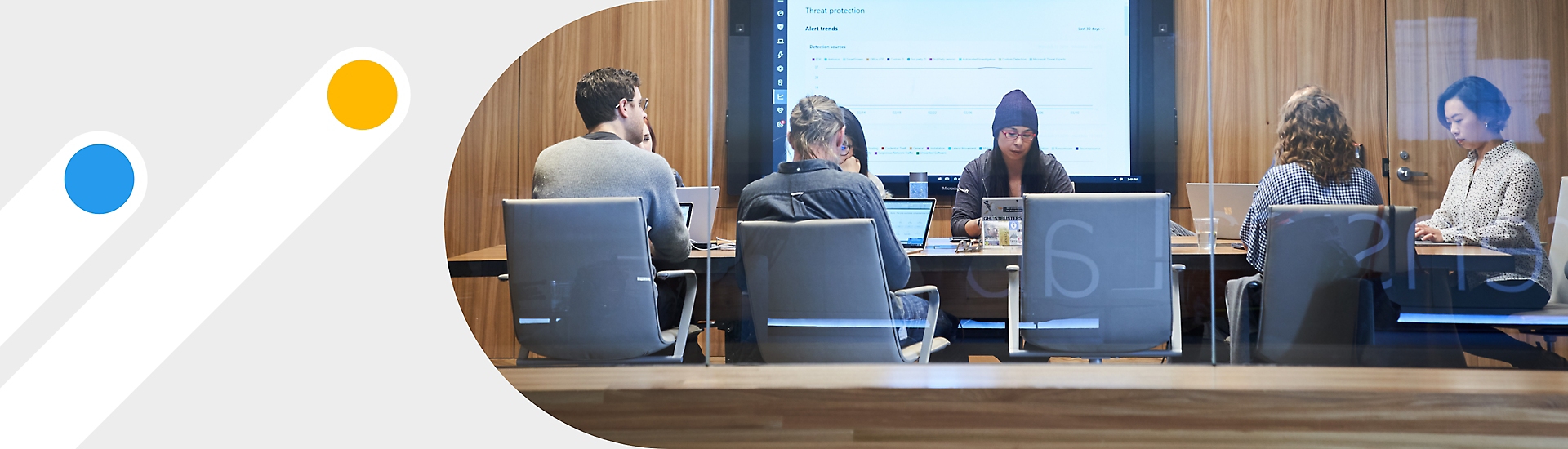 Eine Besprechung mit sieben Personen in einem Konferenzraum, an dessen Rückwand ein Großbildschirm mit Notizen in Teams dargestellt ist