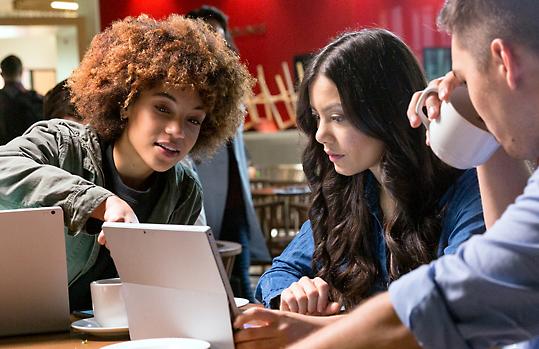 Kolme henkilöä istuu kahvilassa Surface Bookia ja Surface Prota käyttäen. Yksi henkilö osoittaa Surface Pron kuvaruutua kahden muun katsoessa.