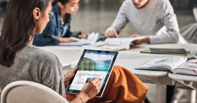 Foto kolmest inimesest, kes istuvad laua taga. Kaks neist loevad koos raamatuid, teine aga kasutab tahvelarvuti ekraanil dokumendi redigeerimiseks pliiatsit.