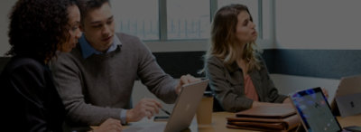 Three people seated at a table with multiple laptops and devices. Two are looking at a laptop. One is watching something out of frame.