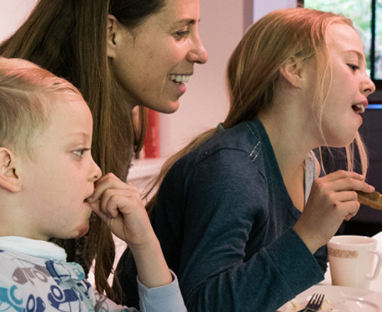 An adult and two children in the kitchen having breakfast and using a table to have a video call with a deployed relative