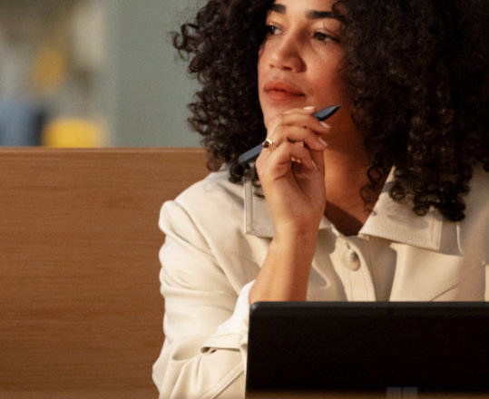 Personne assise seule devant un ordinateur portable, tenant un stylo et regardant pensivement sur le côté.