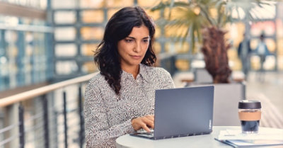 Photograph of a person sitting at a table in an atrium working on a laptop. There are people walking in the background.