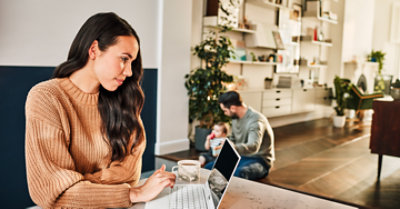 Photograph of family of three at home. One parent is sitting on floor holding young child while other parent sits at table typing on a laptop.