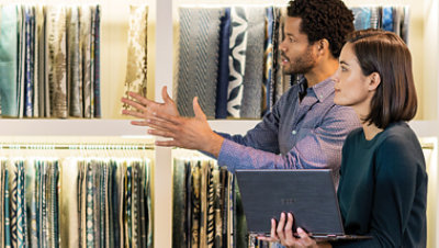 Dans un magasin, un homme et une femme discutent en regardant les tissus exposés. La femme tient un ordinateur portable.