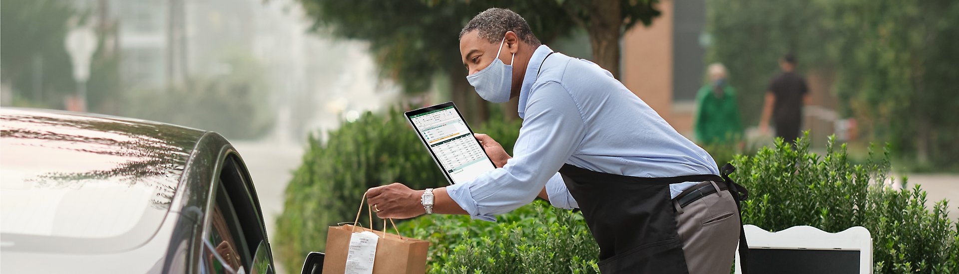 A worker holding a tablet device while leaning over to speak to someone in a car.