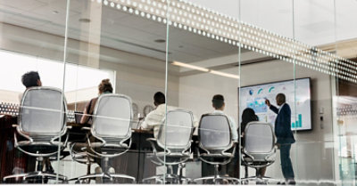 A meeting in a conference room with a presenter pointing at a large monitor displaying data visualizations