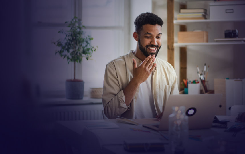 Person engaged in a video call on a laptop at a home office desk, smiling and gesturing using American Sign Language.