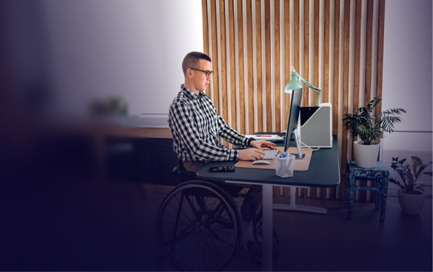 A person who uses a wheelchair works on a desktop computer at a height-adjustable desk in a modern, plant-filled office.