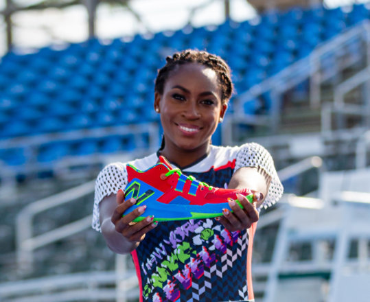 Tennis player, Coco Gauff, smiling and holding up the brightly colored New Balance tennis shoe she designed.