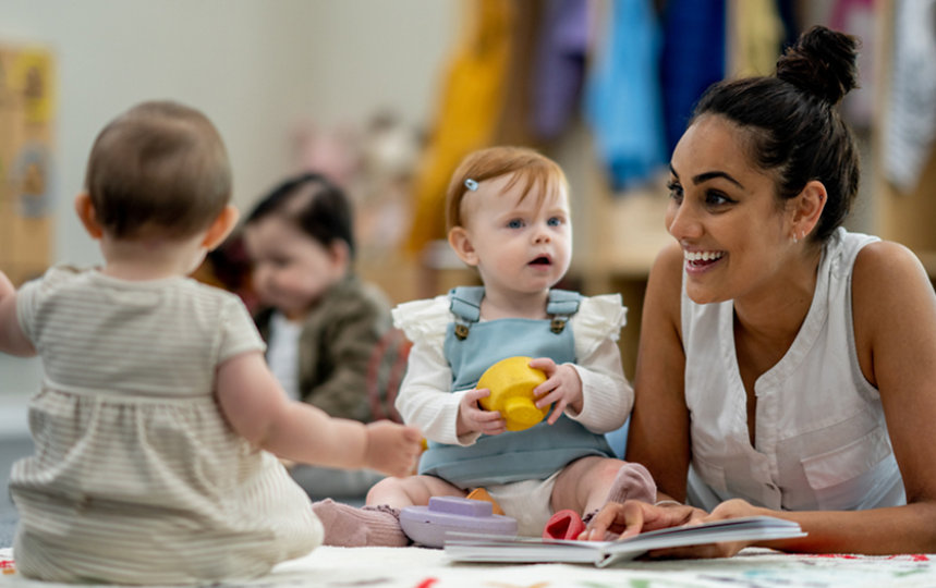 A caretaker smiles as she reads a picture book to two babies.