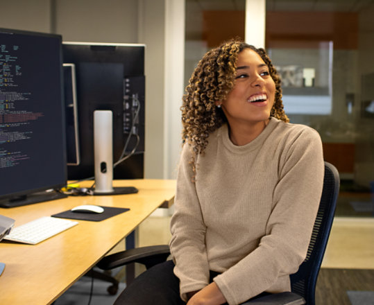 A young Black woman turns away from her computer where she is writing code and smiles.