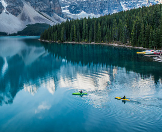 Two kayakers paddle across a calm lake that reflects snow-capped mountains.