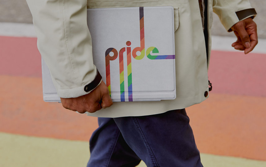 Close up of a person in a trench coat walking with a laptop in a Pride type cover tucked under his arm.