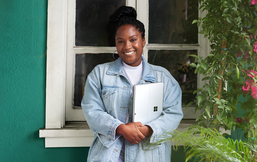 A casually-dressed woman stands outside a stunning green stucco building holding a Surface Laptop and smiling at the camera.