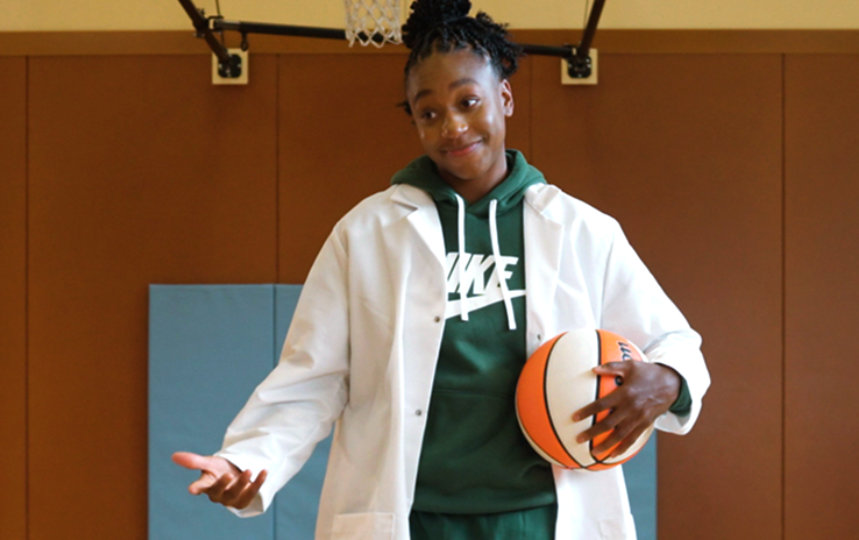 WNBA All Star, Jewell Lloyd, wears a lab coat and holds a basketball as she gives a presentation on STEM and basketball.