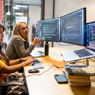 Two women working and discussing with multiple monitors on the desk