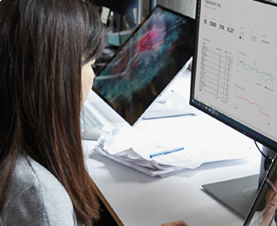 A person on a Teams video call on a laptop and two monitors displaying data and reporting.