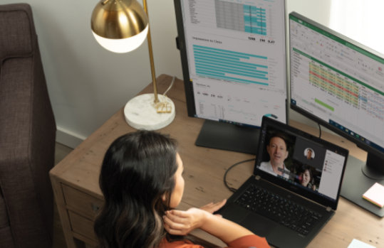 Una persona sentada en un escritorio participando en una videollamada de Teams en un portátil frente a dos monitores.