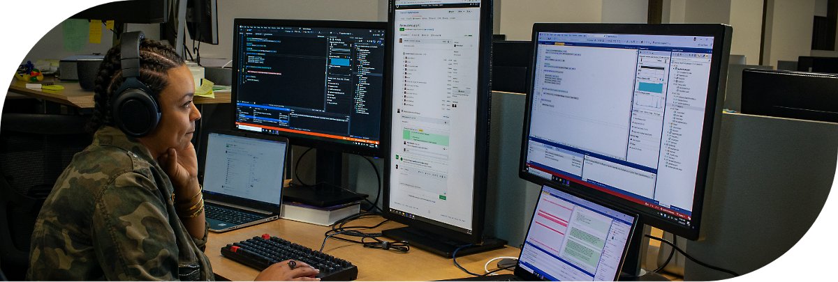 En person med hovedtelefoner, der arbejder ved et skrivebord, hvor der står to bærbare computere og flere skærme.