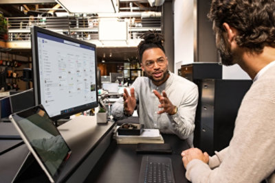 Two people having a conversation at a desk where Teams is being displayed on the monitor.